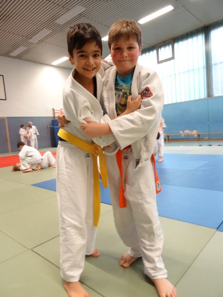 2 Jungs in Judo-Anzügen halten sich, im Hintergrund üben weitere Jungs